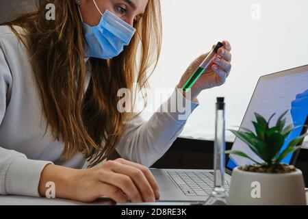 La scienziata femminile sta guardando su un tubo di laboratorio. Matracci con liquido verde e blu vicino alla pianta. Ricerca la cura del virus Foto Stock