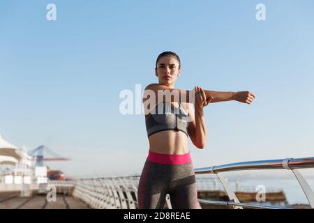 Immagine di bella donna sana in abbigliamento fitness che stretching il suo corpo prima di fare jogging formazione. Corridore femminile in piedi sul lungomare Foto Stock