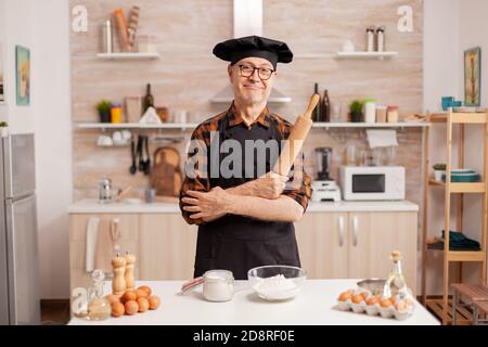 Uomo anziano che indossa lo chef bonete sorridendo in cucina casalinga. Panettiere pensionato in cucina uniforme preparare ingredienti di pasticceria su tavola di legno pronti a cucinare pane, torte e pasta gustosi fatti in casa Foto Stock