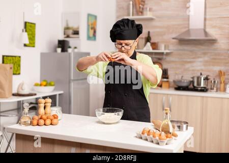 Una donna prepara un impasto per la cottura delle uova cracking in cucina casalinga. Anziano pasticciere spezzare l'uovo su una ciotola di vetro per la ricetta della torta in cucina, mescolando a mano, impastando gli ingredienti prreparing torta fatta in casa Foto Stock