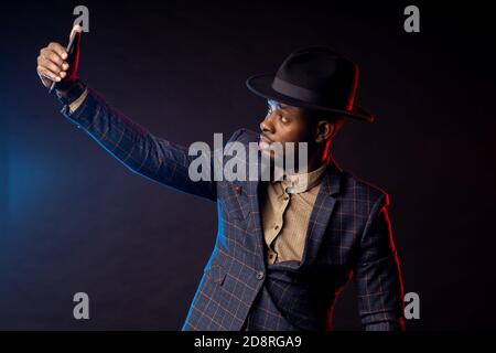 Un giovane afroamericano alla moda con bearded man che indossa un elegante abito a giacca, camicia, cappello, telefono cellulare, che porta selfie al buio, isolato in piedi Foto Stock