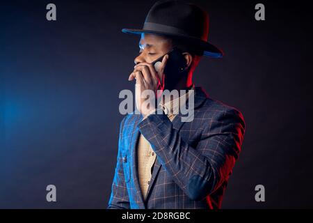Scatto orizzontale di serio preoccupato giovane africano elegante vestito, cappello, parlando sul telefono cellulare, in piedi in buio su sfondo nero . Suc Foto Stock