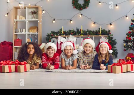 Gruppo di bambini felici a Santa cappelli che guardano la macchina fotografica sdraiato su un piano in un accogliente soggiorno con regali Foto Stock