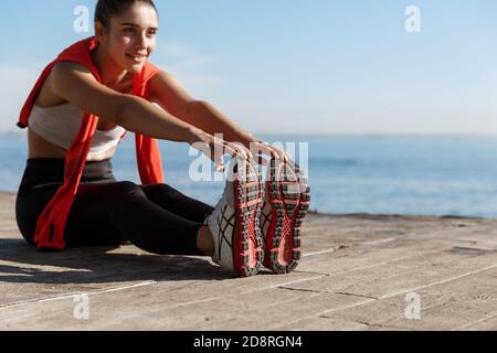 Tiro all'aperto di sorridente sicuro sportivo allenatore vicino al mare. Donna fitness seduta su un molo di legno e gambe che si allungano Foto Stock