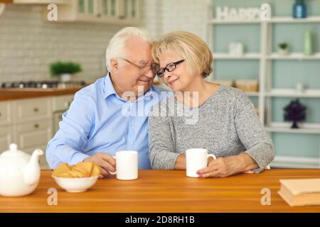 Felice coppia anziana che beve tè e si abbracciano seduti a tavola in cucina Foto Stock