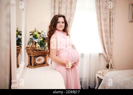 donna incinta dai capelli rossi in un abito rosa in posa in un interno classico vicino alla finestra. in attesa di un bambino. gravidanza Foto Stock