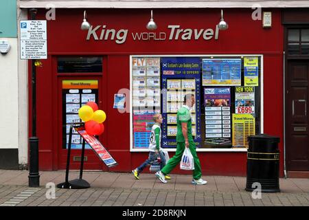 Ayr, Ayrshire, Scozia, Regno Unito. Fotografia di strada candida intorno ad Ayr. Padre e figlio in top calcistici celtici passano da un'agenzia di viaggi dai colori brillanti Foto Stock