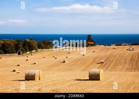 Vista su campo oro del round di balle di paglia alla chiesa del villaggio sulla costa orientale. St Abbs, Berwickshire, Scottish Borders, Scozia, Regno Unito, Gran Bretagna Foto Stock