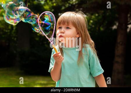 Bambino piccolo, ragazza che soffia bolle enormi da sola, ritratto all'aperto. Bambino giovane con guance soffiato soffia grandi bolle fuori, colpo closeup, ragazza attiva Foto Stock