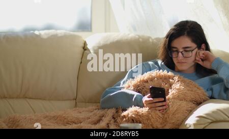 Cute ragazza in occhiali legge le notizie nel suo telefono cellulare, si siede sul divano, sfondo finestra Foto Stock