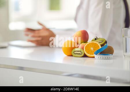 Frutta appetitosa e nastro di misurazione sulla scrivania contro il dietista offuscato dare consiglio di perdita di peso Foto Stock