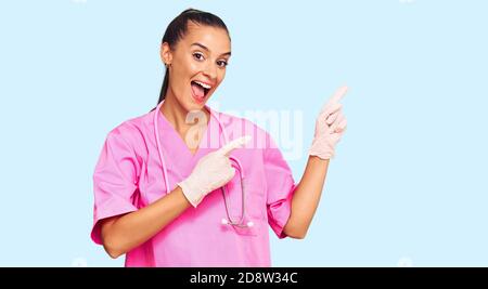 Giovane donna ispanica che indossa uniforme medica e stetoscopio sorridente e guardando la fotocamera che punta con due mani e dita di lato. Foto Stock