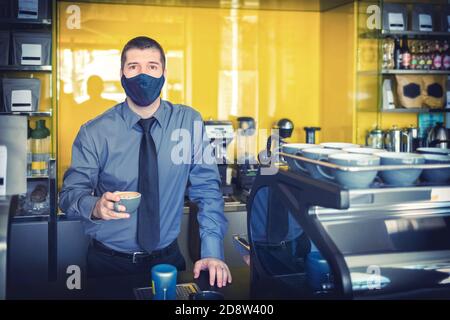 Buon proprietario di piccola impresa con maschera protettiva che serve il caffè dietro il bancone dopo la riapertura del negozio Foto Stock