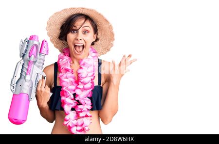 Bella giovane donna con capelli corti indossando bikini e hawaiano lei tenendo pistola ad acqua celebrando la vittoria con felice sorriso e. espressione vincitore wit Foto Stock