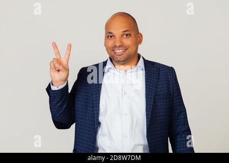 Ritratto di un giovane uomo d'affari afro-americano di successo, mostrando con le dita al numero due, sorridente, fiducioso e felice. L'uomo mostra due dita. Numero 2. In piedi su uno sfondo grigio Foto Stock