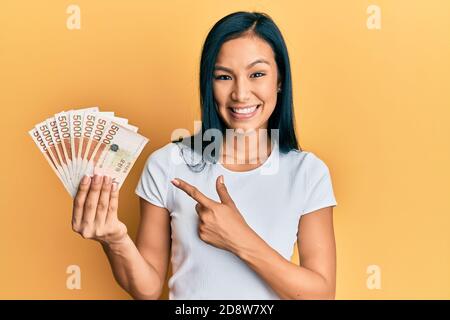 Bella donna ispanica che tiene 5000 sud coreano ha vinto le banconote sorridendo buon puntamento con mano e dito Foto Stock