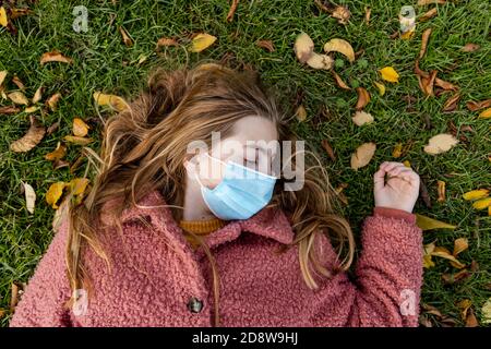 Concetto di ragazza teen bionda che posa sulla sua schiena in erba con una maschera facciale e i suoi occhi chiusi. Stanco del virus Covid. Foto Stock