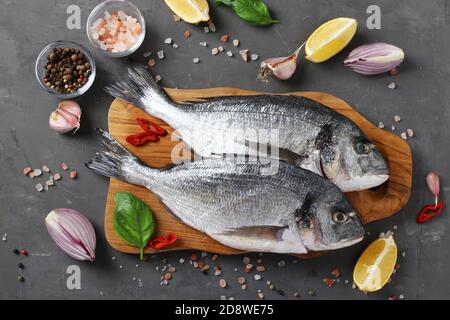 Pesce fresco pronto a cucinare dorado crudo con ingredienti e condimenti come basilico, limone, sale, pepe, cipolla rossa e aglio su tavola di legno al buio Foto Stock