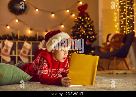 Il ragazzo piccolo apre il libro magico e legge le storie meravigliose e. Favole a casa la vigilia di Natale Foto Stock