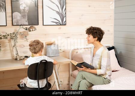 Giovane donna di brunette casual con libro aperto seduto sul letto da suo figlio piccolo Foto Stock