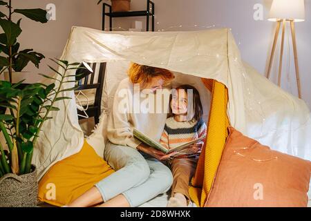 La madre e la figlia giocosa che siedono in tenda fatta di fogli, libro di lettura a casa Foto Stock