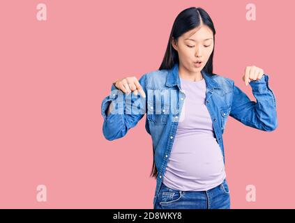 Giovane bella donna cinese incinta in attesa di bambino puntando con le dita che mostrano pubblicità, viso sorpreso e bocca aperta Foto Stock