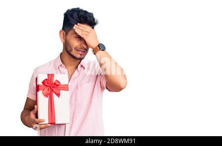 Bell'uomo latino-americano giovane che tiene dono stressato e frustrato con mano sulla testa, sorpreso e faccia arrabbiata Foto Stock