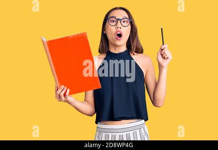 Giovane bella ragazza cinese che tiene il libro spaventato e stupito con bocca aperta per sorpresa, faccia incredula Foto Stock