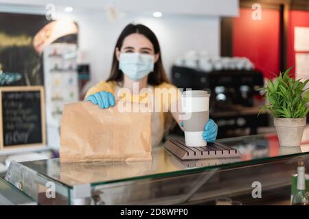Giovane donna che serve caffè da asporto e colazione presso la caffetteria Con maschera protettiva - mettere a fuoco sulla mano destra Foto Stock