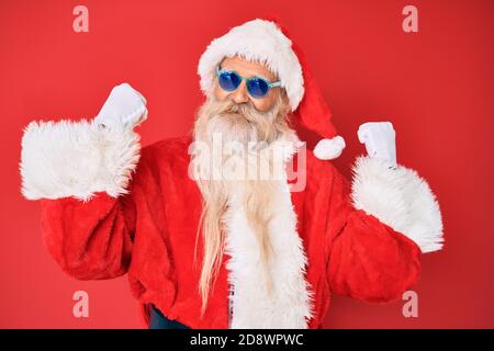 Vecchio uomo anziano che indossa costume di babbo natale e occhiali da sole gridando orgoglioso, celebrando la vittoria e il successo molto eccitato con le braccia sollevate Foto Stock