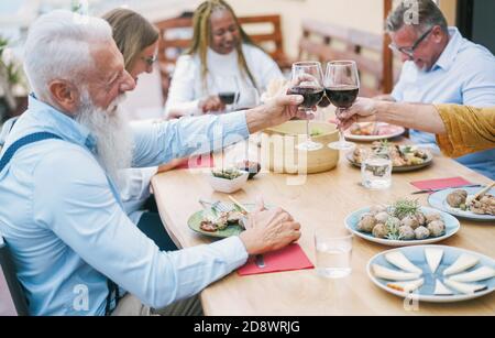 Gente anziana felice che si diverte a cena nel patio - multirazziale Amici che mangiano al pasto di domenica - concentrarsi sul rallegrarsi delle mani con vino Foto Stock