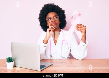 Giovane donna afroamericana che indossa uniforme medico che tiene il nastro di cancro rosa faccia seria che pensa alla domanda con la mano sul mento, pensieroso abou Foto Stock
