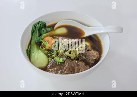 Vista del fuoco selezionata alla zuppa di noodle di manzo allo stufato taiwanese senza noodle in ciotola bianca sul tavolo bianco. Foto Stock