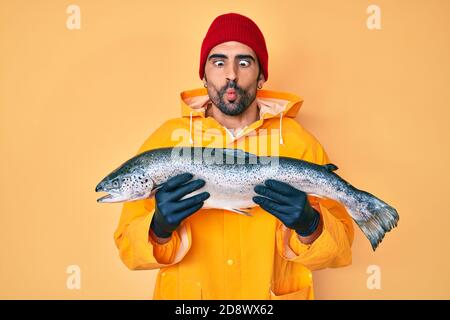 Bell'uomo ispanico con barba che indossa attrezzature da pesca facendo viso di pesce con bocca e occhi squinting, pazzo e comico. Foto Stock