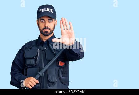 Giovane uomo ispanico che indossa uniforme di polizia tenuta baton con mano aperta facendo stop segno con espressione seria e sicura, gesto di difesa Foto Stock