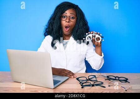 Bella donna africana che tiene occhiali di optometria spaventato e stupito con bocca aperta per sorpresa, viso incredulo Foto Stock