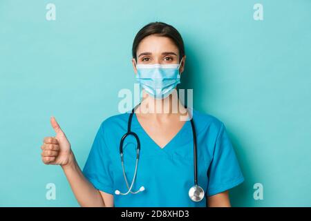 Concetto di covid-19 e concetto di quarantena. Primo piano di sicuro medico femminile in maschera medica e scrub, tenendo stetoscopio, mostrando pollici-up Foto Stock
