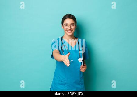 Concetto di coronavirus, pandemia e allontanamento sociale. Immagine di una donna sorridente in scrub, tenendo appunti, allungare la mano per la stretta di mano Foto Stock