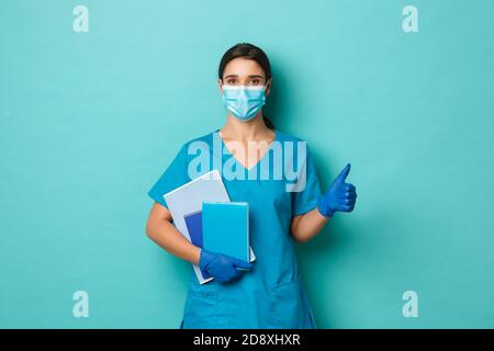 Covid-19, pandemia e concetto di medicina. Immagine di un medico sicuro in maschera medica, guanti e scrub, tenendo taccuini e mostrando pollici-up Foto Stock
