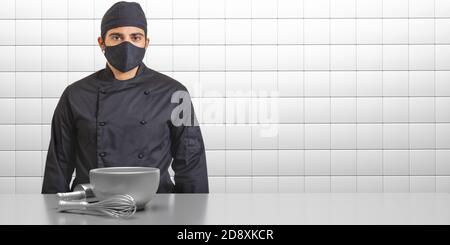 Chef nei giorni del coronavirus. Uomo in uniforme cuoco con maschera facciale nera, fondo interno cucina. COVID19 sicurezza nella preparazione di alimenti per ristoranti Foto Stock