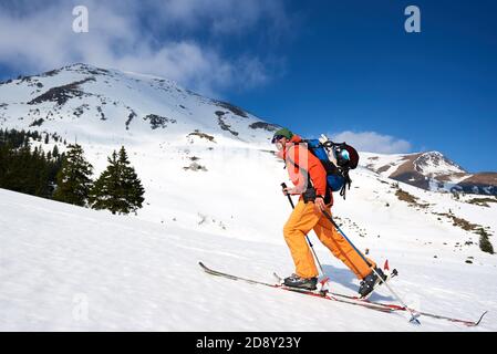 Sciatore turista zaino in spalla, felice sorridente uomo bearded trekking sugli sci su collina sullo sfondo del cielo blu e belle creste di montagna. Vacanze invernali, stile di vita attivo, concetto di sci e trekking. Foto Stock