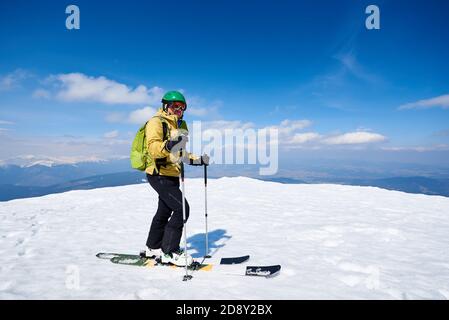 Sciatore sportivo con zaino in casco e occhiali in piedi sugli sci sulla cima della montagna innevata su copy space sfondo di cielo azzurro luminoso e paesaggio di altura. Concetto di attrezzatura da sci professionale. Foto Stock