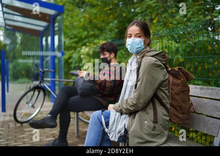 Giovane donna con maschera facciale sulla fermata dell'autobus all'aperto in città. Concetto di coronavirus. Foto Stock