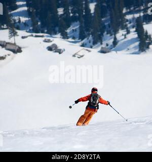 Vista posteriore di sciatore professionista in casco con zaino sci in neve profonda sullo sfondo di villaggio di montagna o case di resort tetti e alberi di abete verde. Vacanze invernali e concetto di ricreazione. Foto Stock