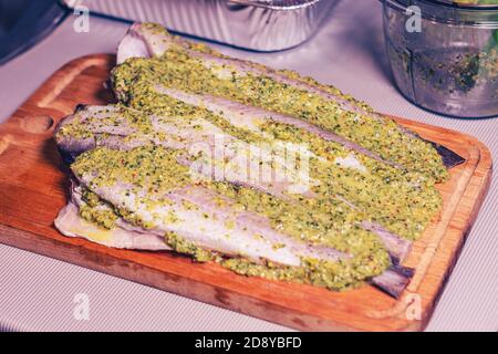 Filetto di pollock crudo con salsa verde su un tagliere di legno in cucina. Primo piano del processo di cottura. Foto Stock