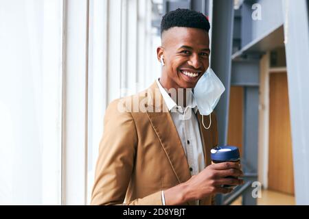 Ritratto di giovane uomo d'affari che indossa la maschera che tiene il caffè in piedi dentro Ufficio moderno durante Pandemic Foto Stock