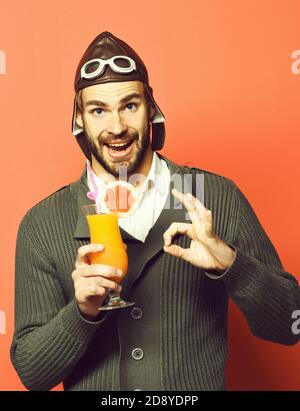 bell'uomo aviatore con bearded e barba lunga sul volto sorridente che tiene un bicchiere di cocktail alcolico in maglia grigia con cappello e bicchieri, su sfondo rosso dello studio Foto Stock