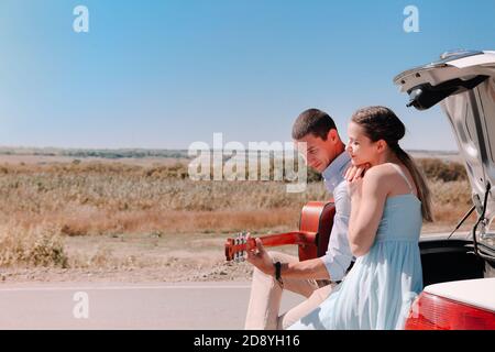 Felice giovane coppia che suona la chitarra e che si gode il tempo insieme mentre seduto sul bagagliaio aperto dell'auto parcheggiata su strada di campagna durante il viaggio estivo Foto Stock