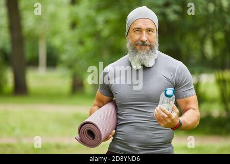 Ritratto medio di uomo anziano con bearded bello che tiene il tappeto di yoga e una bottiglia d'acqua che si trova nel parco guardando la macchina fotografica Foto Stock