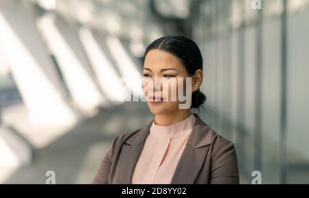 Bella donna d'affari asiatica guarda il lato in piedi sulla strada all'aperto. Brunetta di mezza età con giacca marrone. Sfondo a trama bianca. Foto Stock